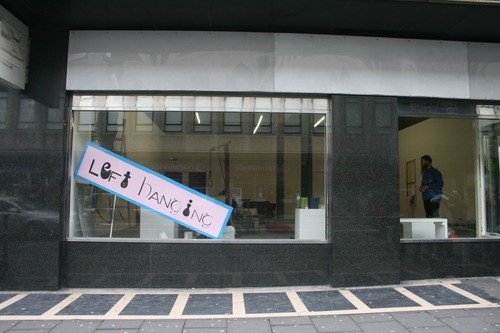 A hand-painted pink sign reading 'Left Hanging' propped in a shopfront window.